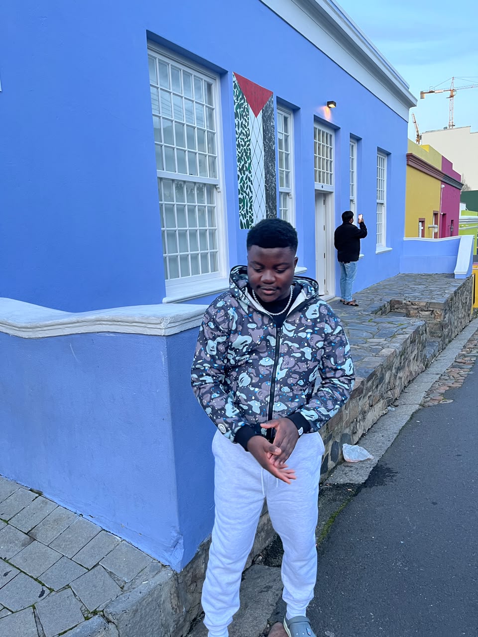 Kupela on a cobbled street in Bo-Kaap, in front of a bright blue house with the quarter's painted houses behind