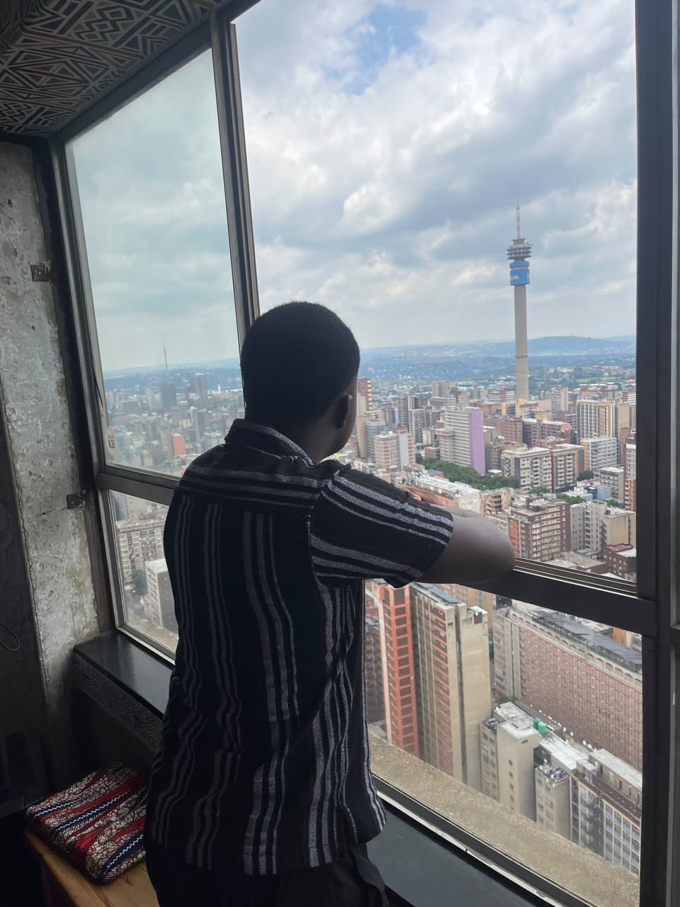 Kupela at a high window in Ponte Tower looking out over the Johannesburg skyline, the Hillbrow Tower on the horizon