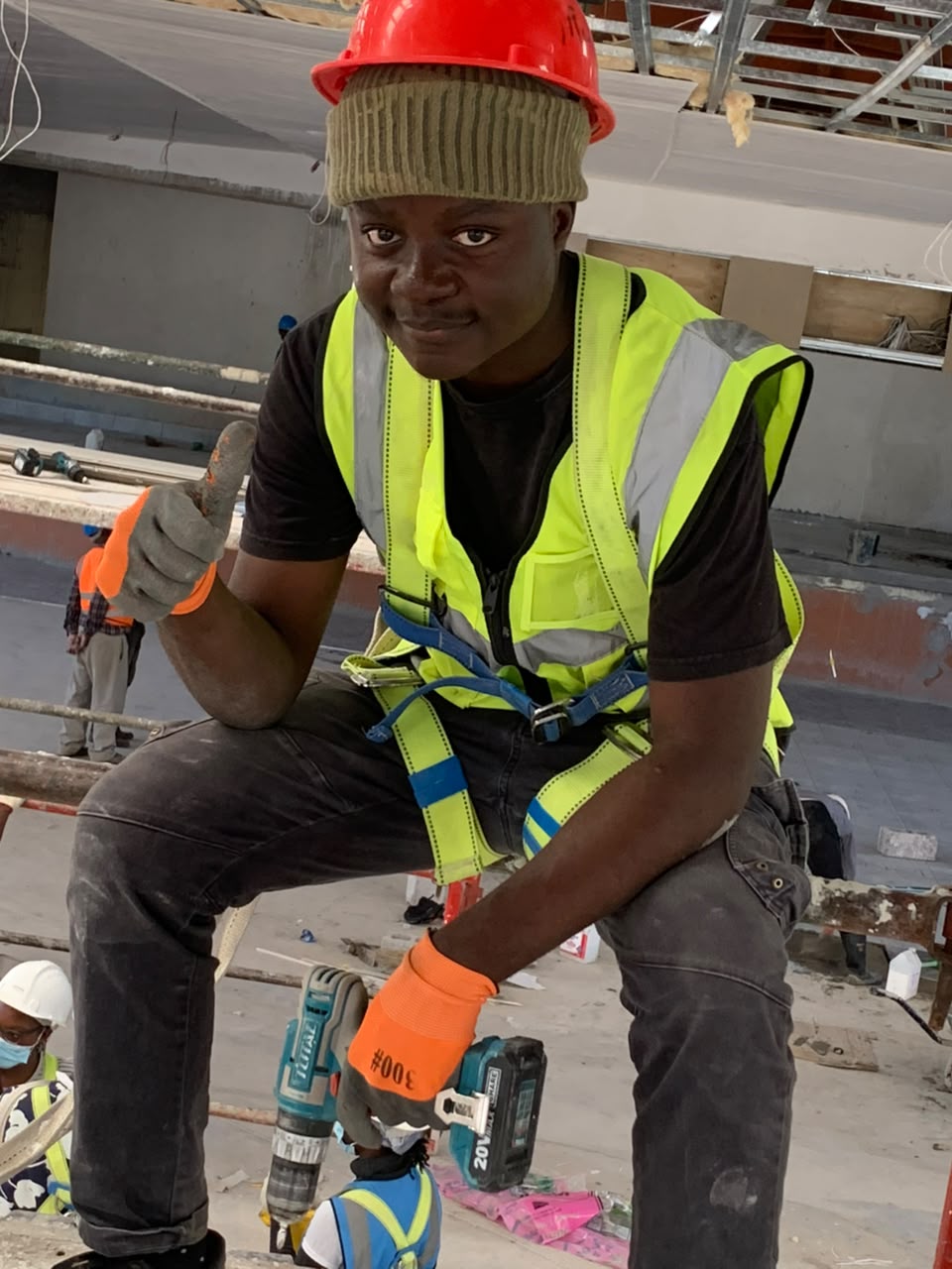 Kupela in a hard hat, hi-vis vest and safety harness on a building site, crouched beside a power drill